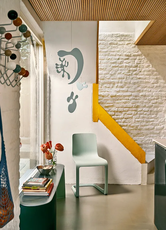 Heller Eingangsbereich mit Vitra Freischwinger-Stuhl in Hellgrün, Tisch mit Büchern, Vase, gelber Treppe, Holzdecke und Garderobenhaken.